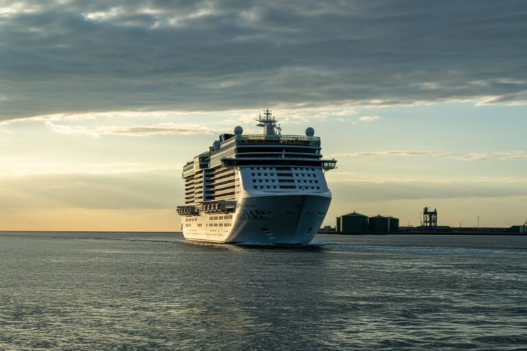 Large white cruise ship near a harbor at sunset, calm water reflecting the sky.