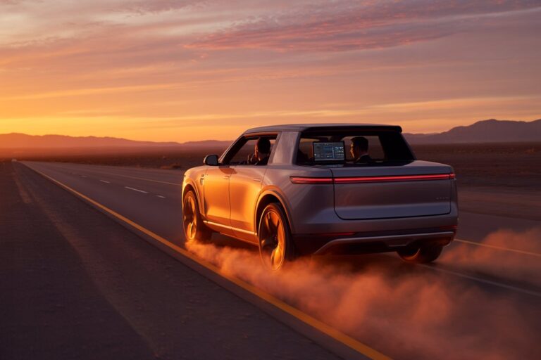 Silver SUV seen from the rear-side, driving on a desert highway at sunset with dust clouds trailing behind.