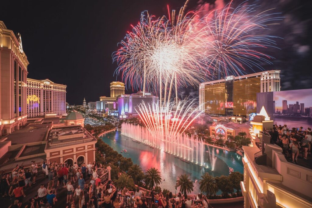 Fireworks brighten the night sky over the Bellagio fountains in Las Vegas as crowds watch the show from the surrounding terraces and palm trees glow below.