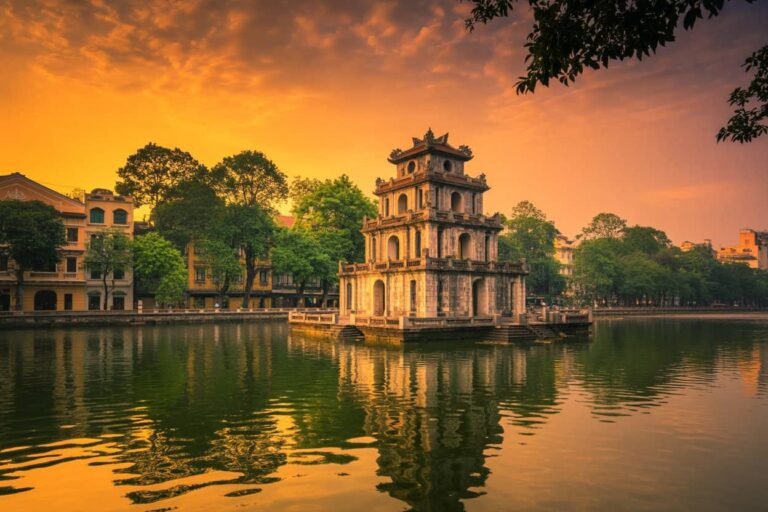 Three-story stone tower on a small island in a lake at sunset, with trees and glowing orange sky reflecting in the water.