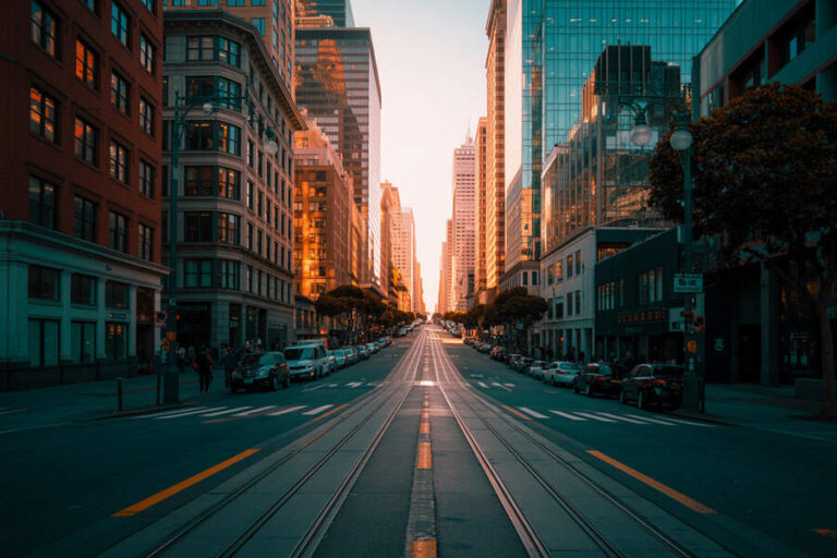 City street with tram tracks in the middle, tall office buildings on both sides, and cars parked along the curb at sunset.