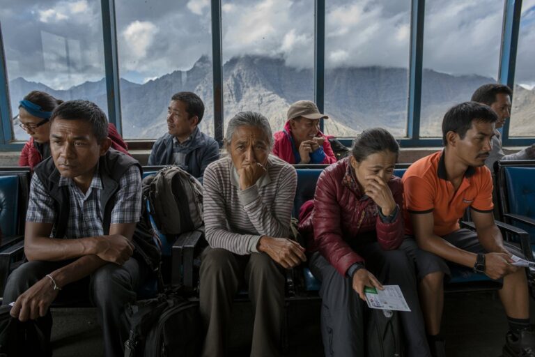 Group of travelers sitting on a bus with backpacks, gazing pensively as mountains rise outside the windows that line the cabin boundary.