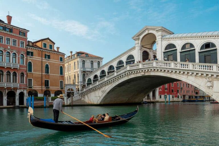 Gondola with passengers gliding under a white arched bridge along a Venetian canal, colorful buildings on the quay in the background.
