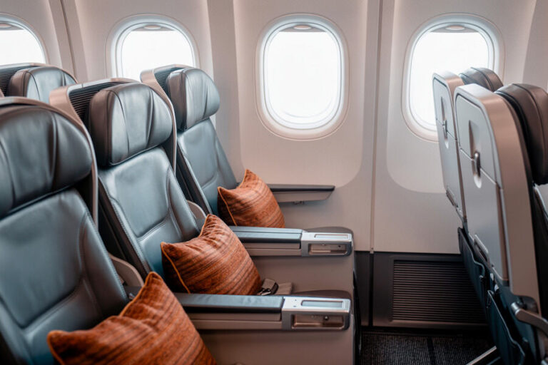 Airline cabin interior with gray leather seats and orange throw pillows by the windows.