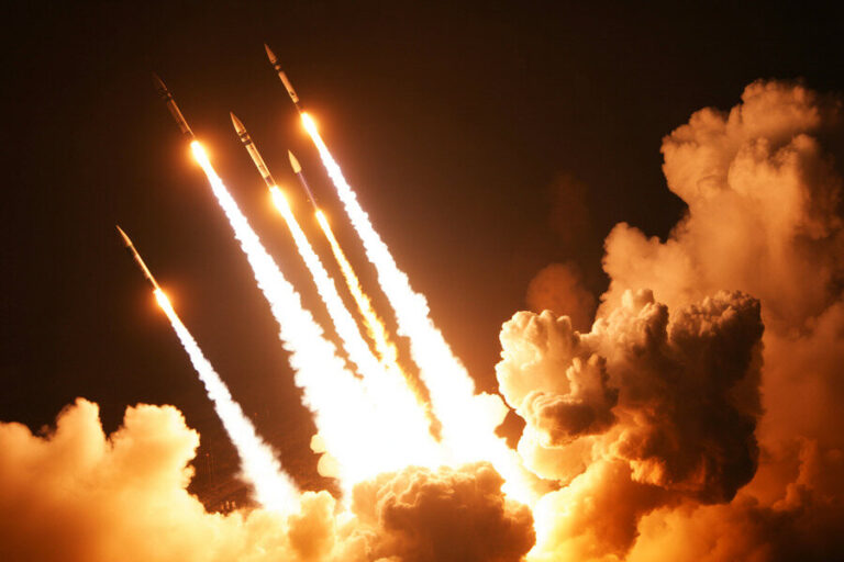 Rockets launch at night, leaving bright flames and smoke plumes against a dark sky.