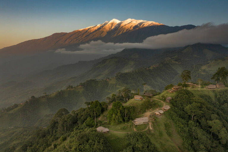 Aerial view of a green hillside village with thatched huts, mist in the valley, and snow-capped mountains at sunrise.
