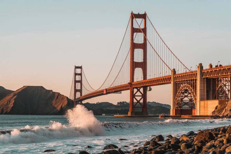 Golden Gate Bridge at sunset, orange towers over a rocky shoreline with waves crashing nearby.