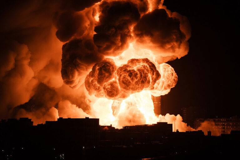 Massive orange fireball and dark smoke rising above a cityscape at night after an industrial explosion.