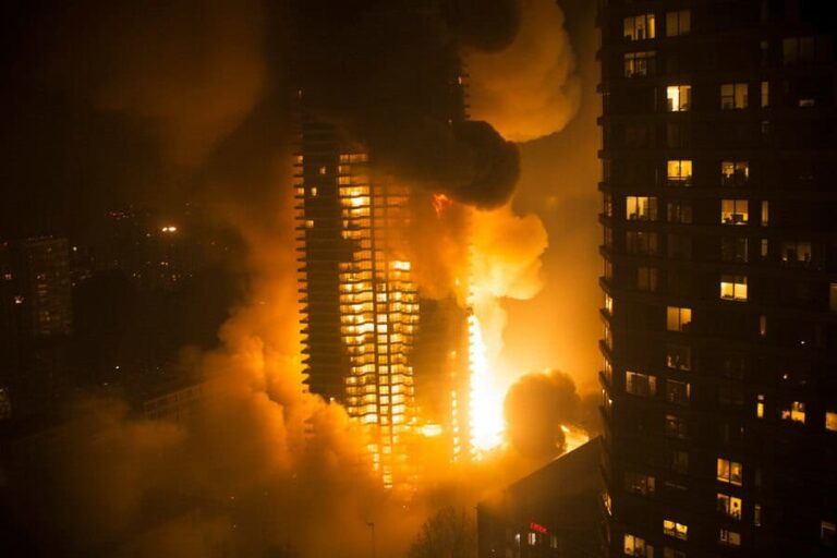 High-rise building on fire at night with bright flames and thick smoke engulfing surrounding towers, city lights dimmed by smoke.