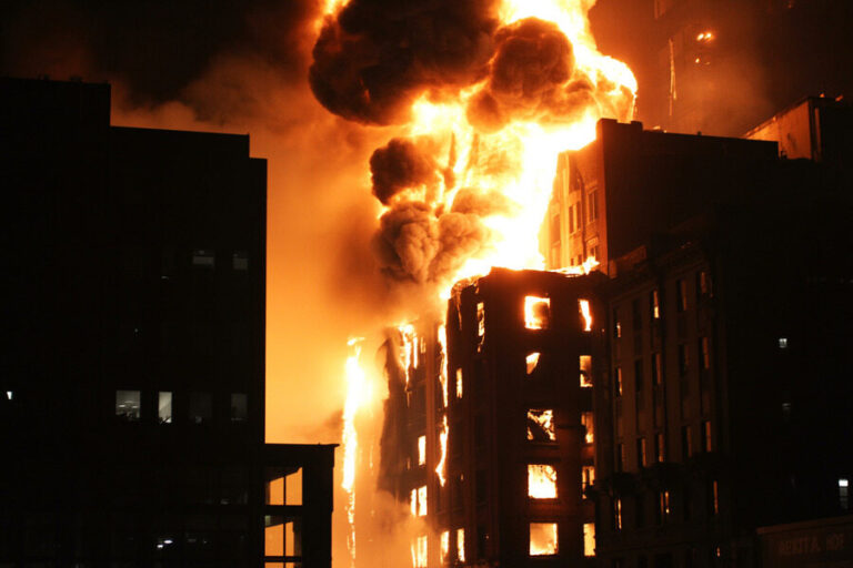 A tall building engulfed in bright flames with billowing black smoke at night, windows glowing from the fire.