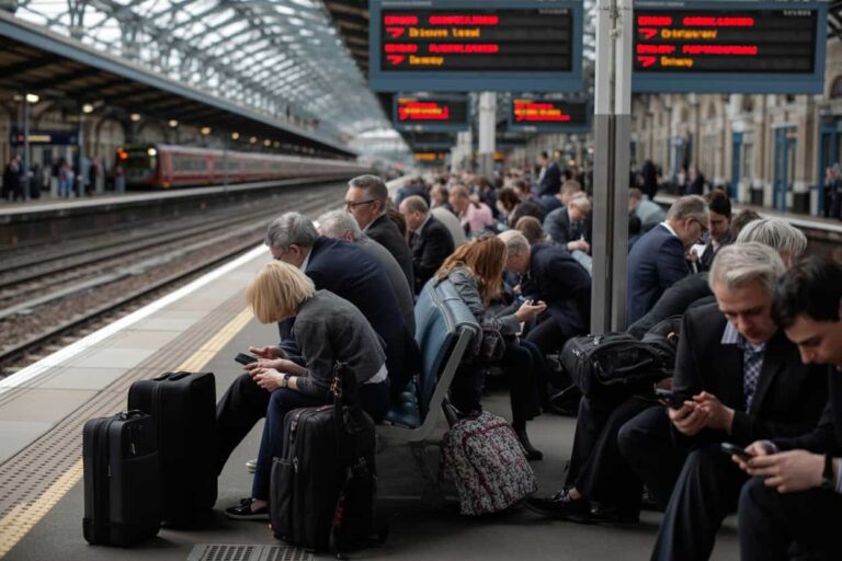 Crowded train platform with travelers seated along the edge, many looking at phones and surrounded by luggage.