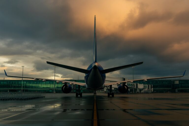 Front view of a large commercial airplane on a wet tarmac at sunset, jet wings outstretched and terminal in background.
