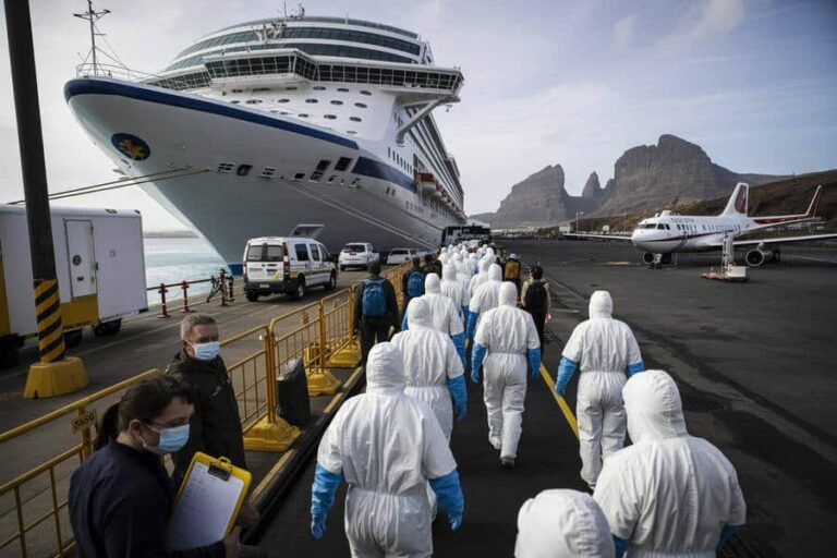Line of people in white protective suits walking along a dock toward a large cruise ship.