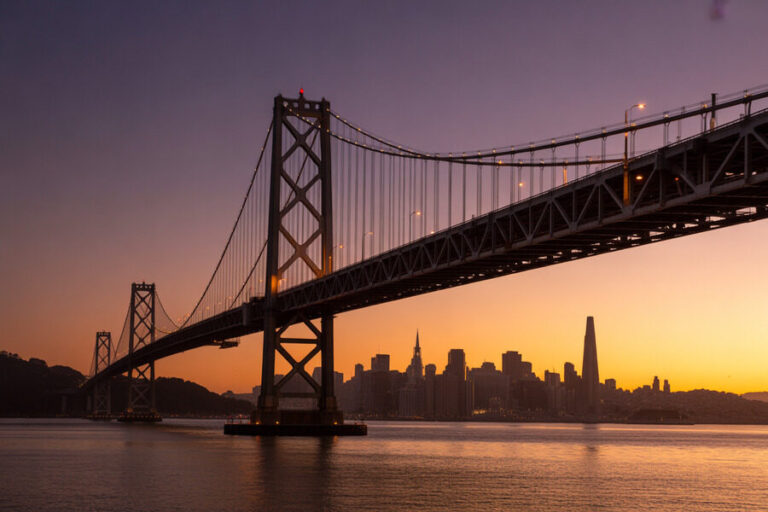 Suspension bridge spanning a calm waterway at dusk, with a warm orange-purple sunset and a distant city skyline silhouetted.