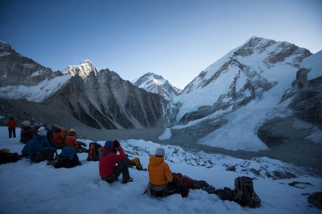 A group of mountaineers resting on a snow-covered slope with colorful jackets and backpacks, as tall, rugged, snow-dusted mountains rise in the background under a clear blue sky.