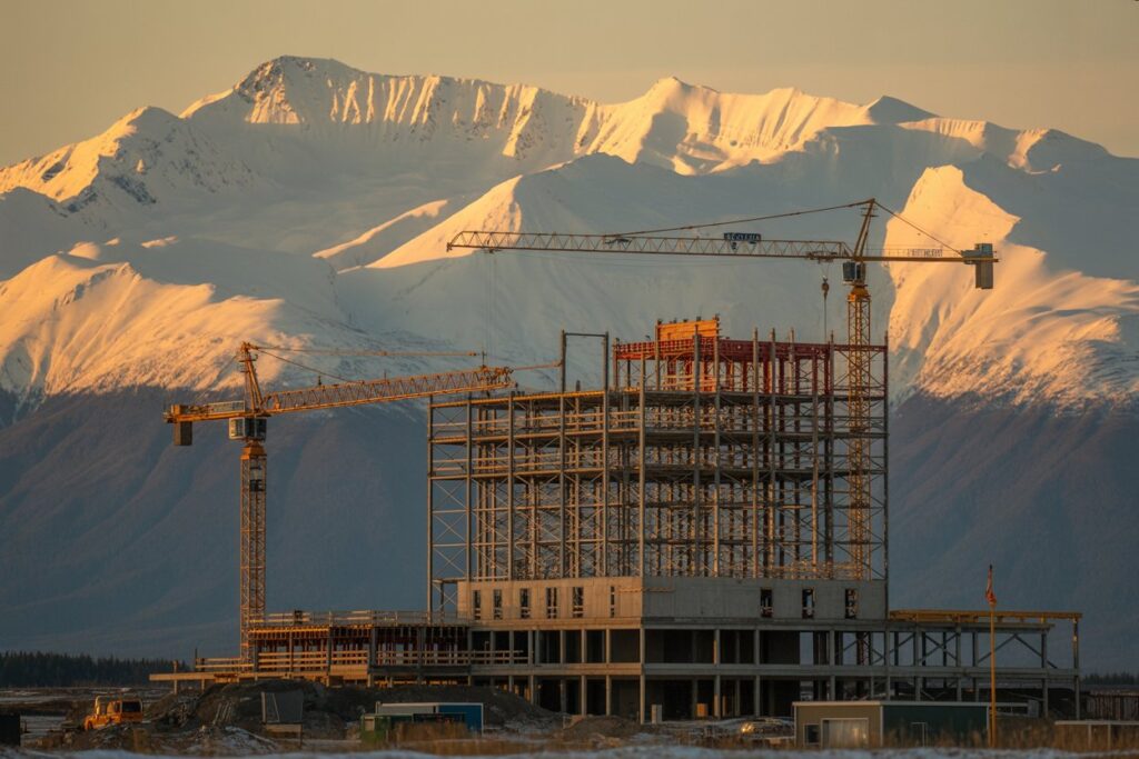 Construction site with a tall steel frame and two cranes set against snow‑capped mountains at sunset