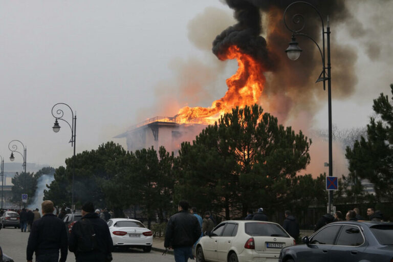 Building on fire with large orange flames and dark smoke rising over trees, while bystanders and parked cars line the street.