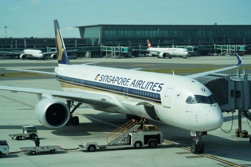 Singapore Airlines jet parked at the gate with a boarding bridge attached and ground support nearby.