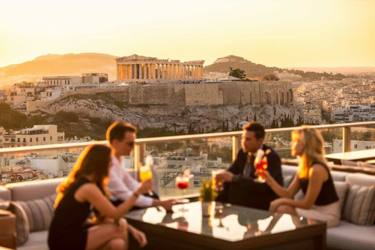 Four people on a rooftop terrace at sunset, chatting and sipping cocktails with the Acropolis in the background.