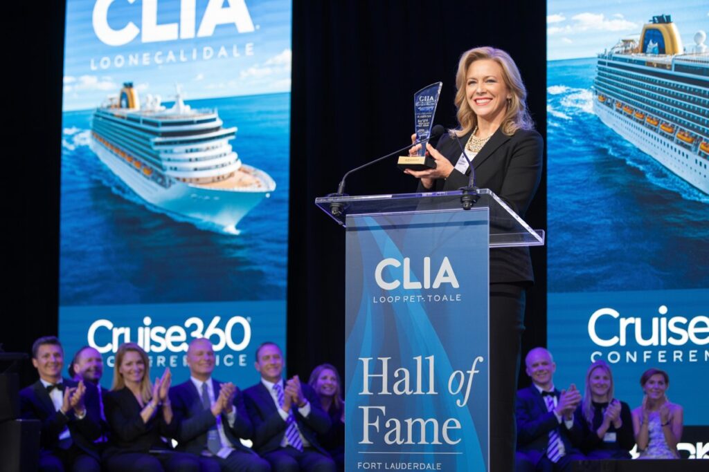 Woman on stage holding an award at a CLIA Hall of Fame event, with cheering group and cruise ship banners in the background.