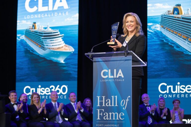 Woman on stage holding an award at a CLIA Hall of Fame event, with cheering group and cruise ship banners in the background.