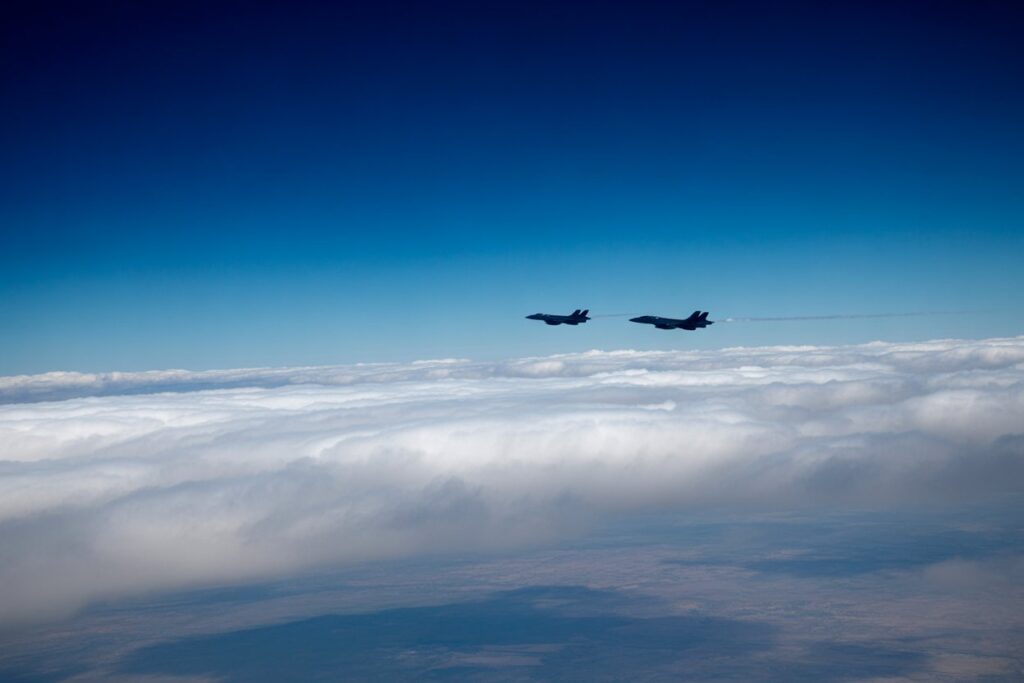 Two military jets fly in formation above a sea of white clouds against a deep blue sky.
