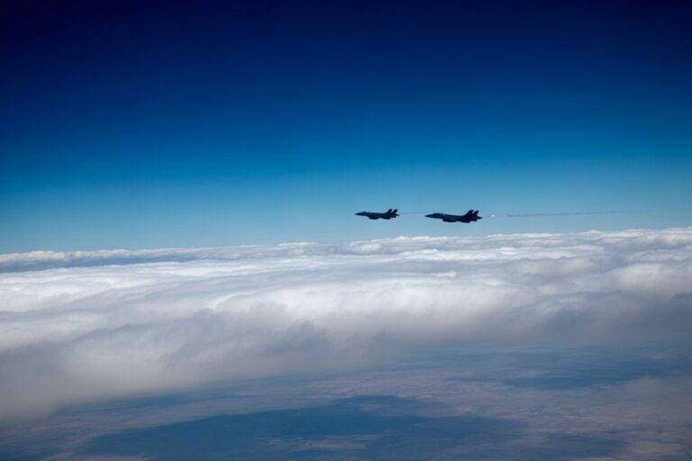 Two military jets fly in formation above a sea of white clouds against a deep blue sky.