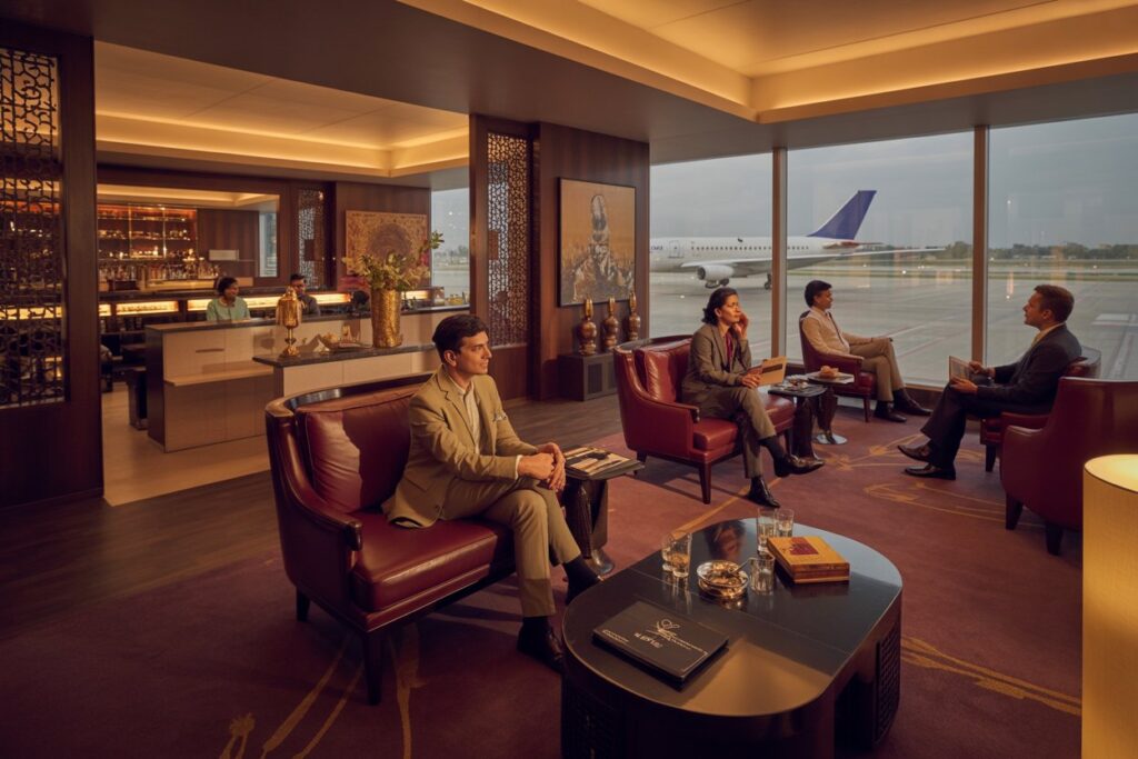 Business lounge at an airport with four suited professionals seated on leather chairs, a bar in the background, and a plane visible through large windows
