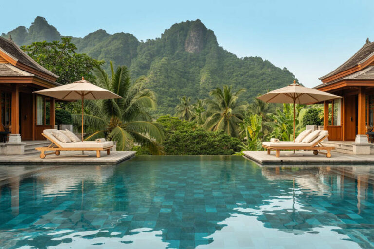 Resort poolside scene with two lounge chairs under umbrellas, wood villas, and lush tropical scenery.