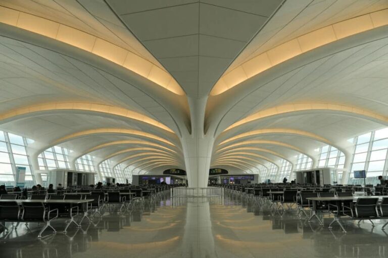 Wide interior of a modern airport terminal with curved white arches, row of black chairs, and large windows left and right.