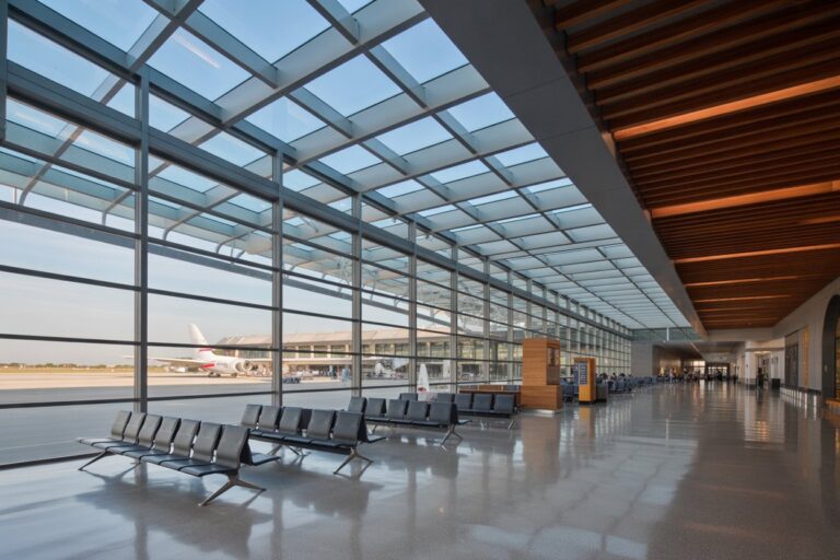 Interior of a modern airport terminal with floor-to-ceiling glass walls, rows of black seats, and a runway view with a parked airplane outside.