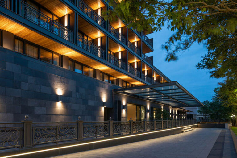 Evening exterior of a modern multi-story building with illuminated balconies and a glass canopy over the entrance.