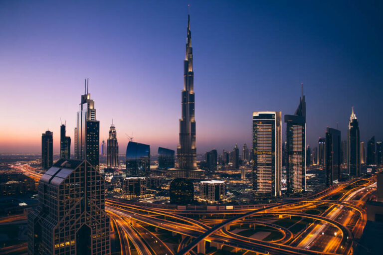 Dubai skyline at twilight with the Burj Khalifa towering above illuminated highways and sleek glass buildings nearby.