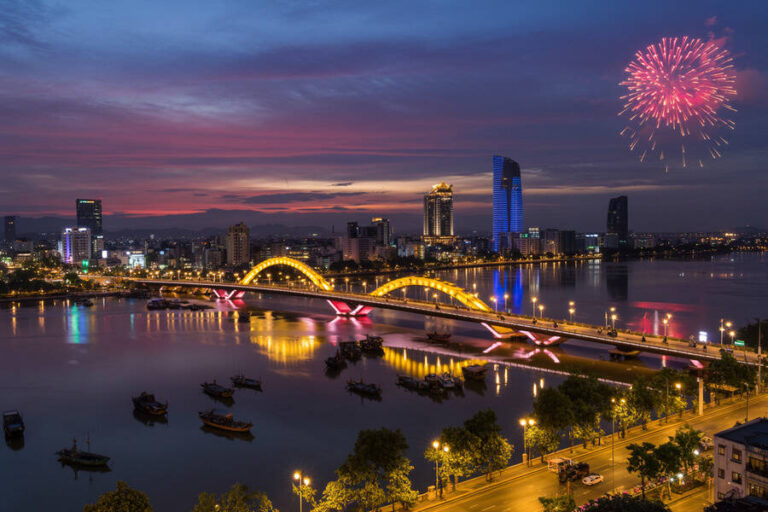Twilight city skyline with a lit arched bridge over a calm river and a fireworks display in the sky above.