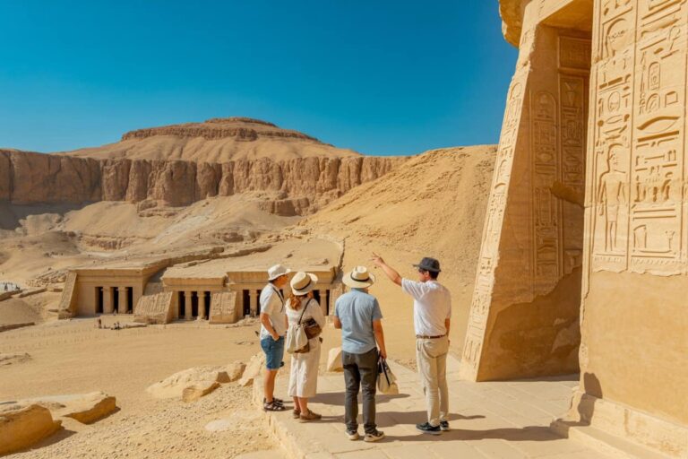 Group of four tourists with hats on a stone platform, listening to a guide beside a hieroglyph-inscribed temple wall in a desert site by blue sky in daylight