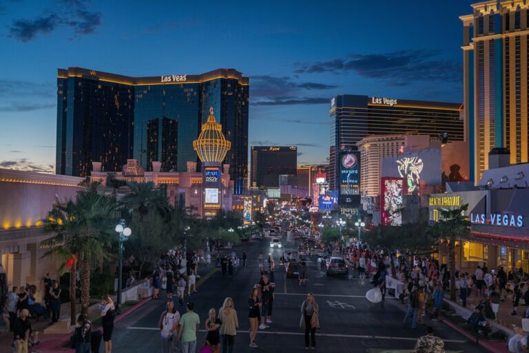 Night view of the Las Vegas Strip with illuminated casinos, palm trees, and pedestrians walking along the street at dusk.