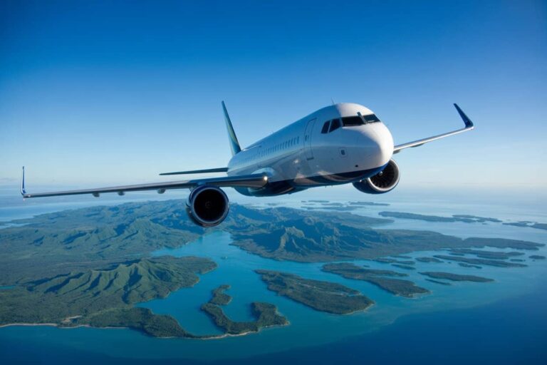 Passenger jet flying over a chain of green islands and blue ocean under a clear sky.