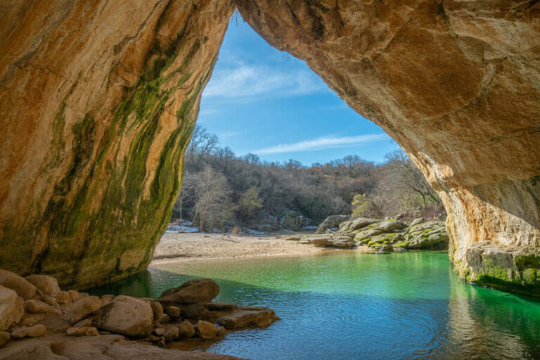 View from inside a rocky cave opening onto a calm turquoise pool and sandy beach beyond under a blue sky with trees in the background.