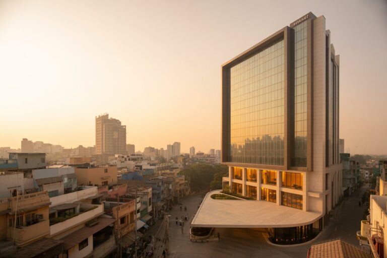 Glass-fronted modern office building reflects the orange sunset, towering over a busy city street with a curved white plaza at its base.