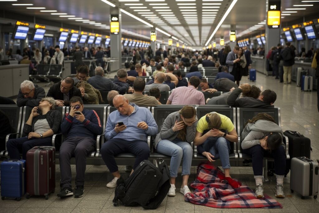 Crowded airport departure hall with travelers seated in rows, many looking at screens or phones, backpacks and suitcases nearby and a red plaid blanket on the floor.|informative image