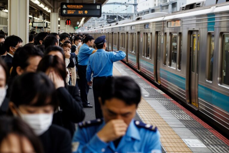 Crowded train platform with masked commuters as a blue-uniform officer waves to a passing silver train.