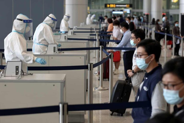 Airport security/check-in area with medical staff in full PPE (P100 suits, masks, face shields) at counters; travelers in line wearing masks with luggage.