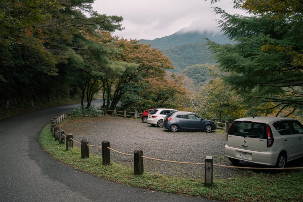 Parking area along a winding mountain road with several cars parked on gravel and a rope fence, surrounded by autumn trees and distant misty hills.