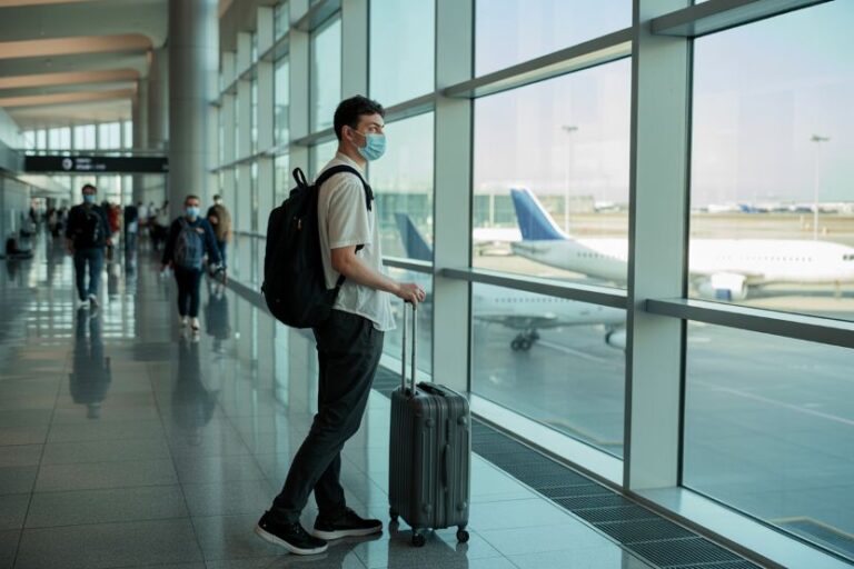 Traveler wearing a mask in an airport terminal with a rolling suitcase, looking toward the windows at parked planes.