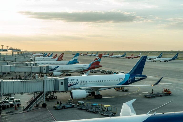 Airplanes parked at a busy airport tarmac with jet bridges and ground vehicles during sunset; multiple gates lined up.