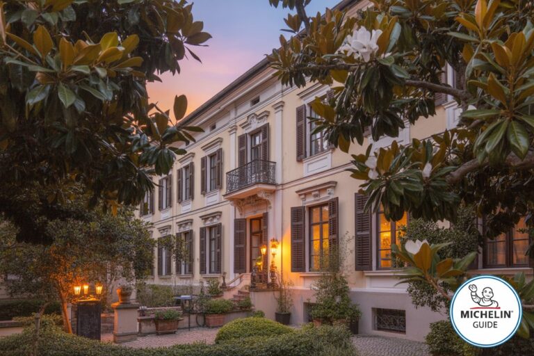Sunset-lit historic villa with cream facade, dark shutters, and a garden courtyard framed by leafy branches.