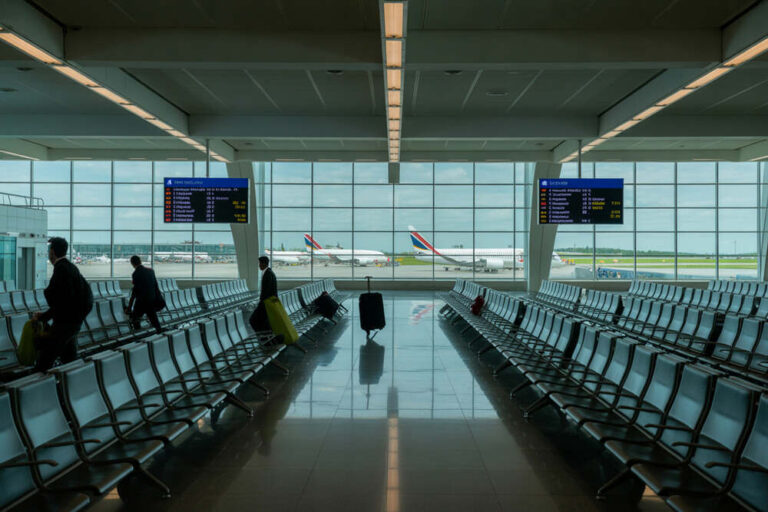 Airport departure lounge with empty and occupied rows of seats, travelers with rolling luggage, and flight boards visible through large windows.