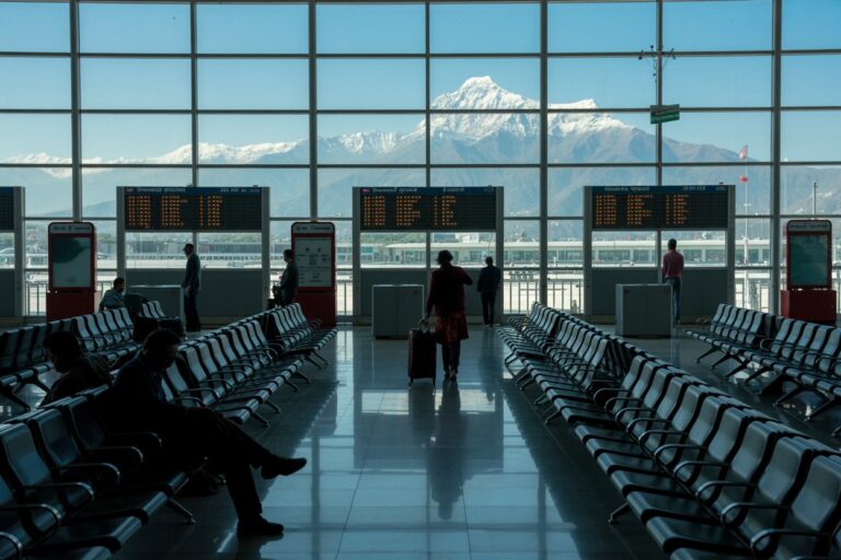 Airport terminal with large glass windows revealing snow-capped mountains outside; travelers sit and wait in rows of chairs nearby.