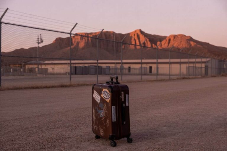 Brown hard-shell suitcase with travel stickers stands on a gravel lot at sunset, with a chain-link fence and desert mountains in the background.
