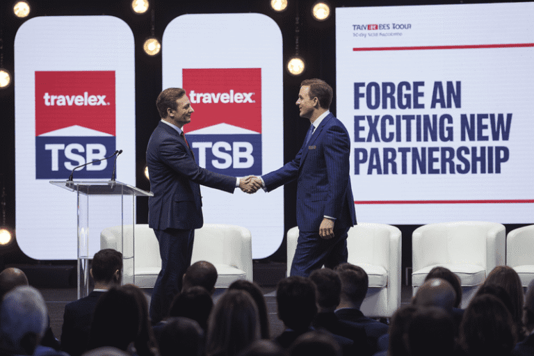 Two men in suits shake hands on a conference stage, behind them screens display Travelex and TSB logos with the words 'Forge an exciting new partnership'.